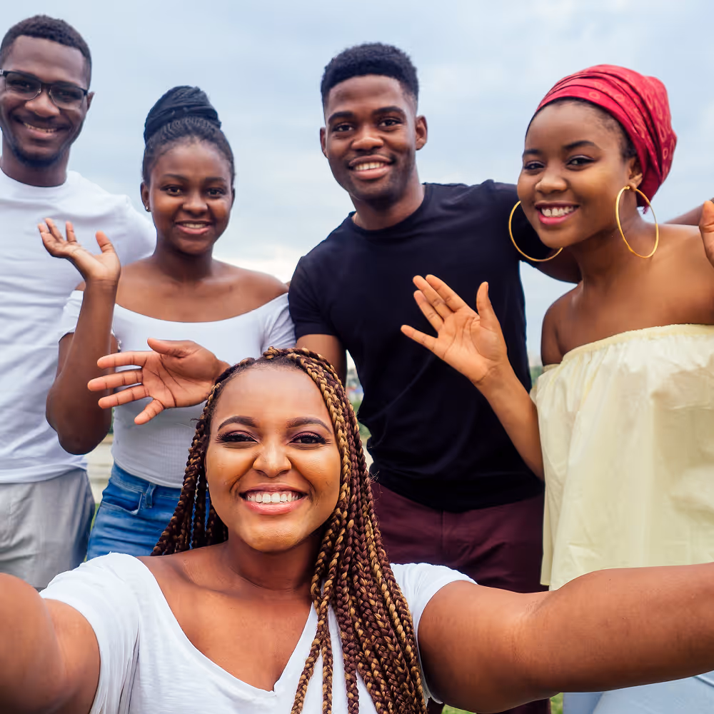 Five young adults smiling and waving at the camera outdoors with a cloudy sky background.